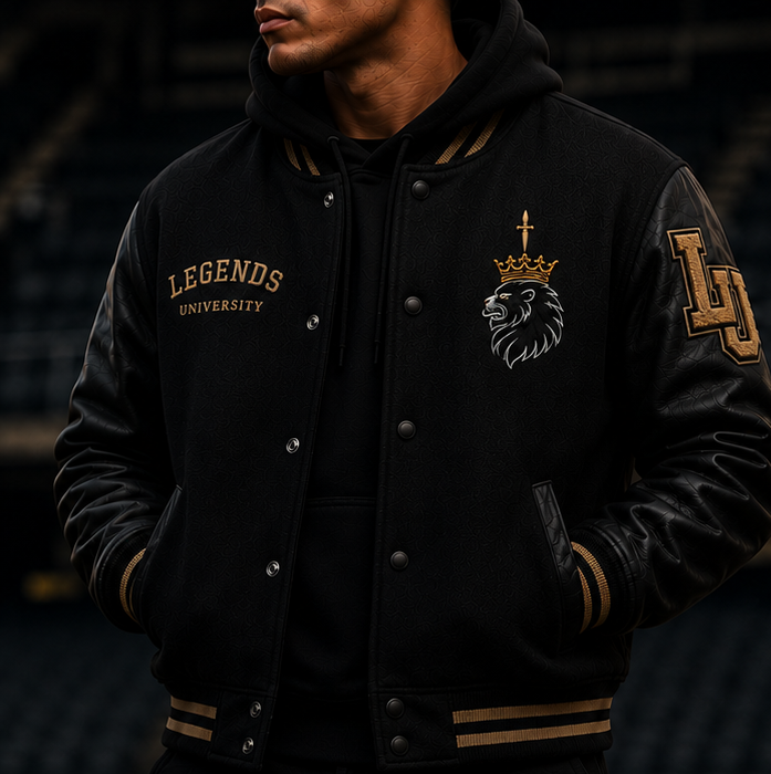 Man wearing a black varsity jacket with black leather sleeves and gold ribbed trim, embroidered with a crowned-lion crest and LU sleeve patch, in a dim arena